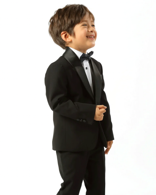 boy wearing black tux, including black shawl lapel blazer, white shirt, black dress pants, and black bow tie. he's standing side on.
