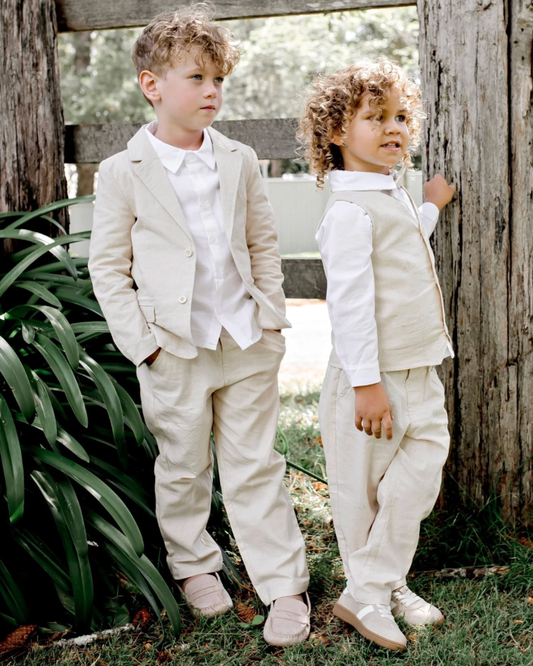 Two young boys wearing matching beige and white formal children's clothing stands outdoors against rustic wooden posts and greenery.