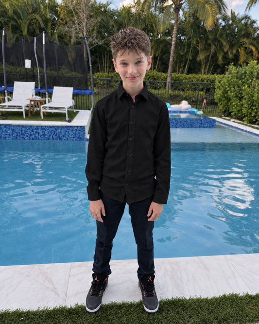 Boy wearing a structured black long-sleeve dress shirt and black trousers beside an outdoor swimming pool.