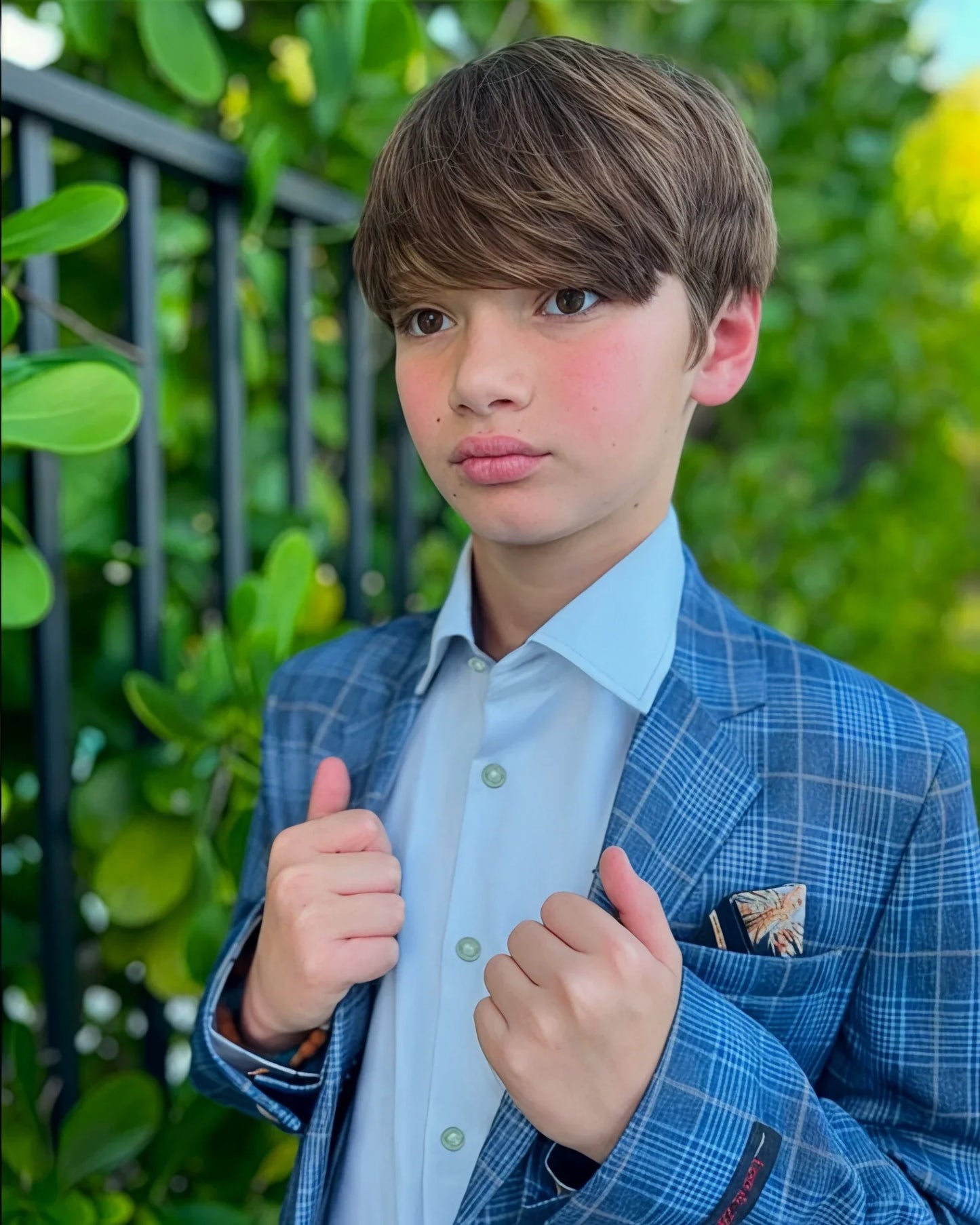 Young boy in a blue checkered suit giving a thumbs-up gesture outdoors.