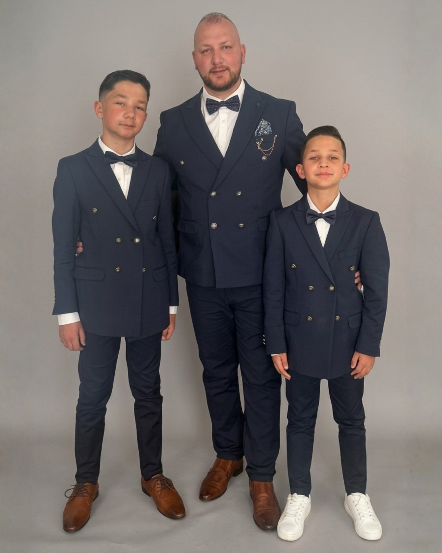 Man and two boys standing together in a studio, wearing matching navy blue double breasted suits with white shirts and bow ties, facing the camera against a plain light grey background