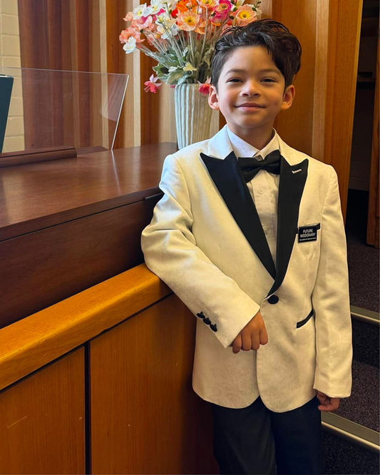 Young boy in a formal white suit with black lapels standing indoors.