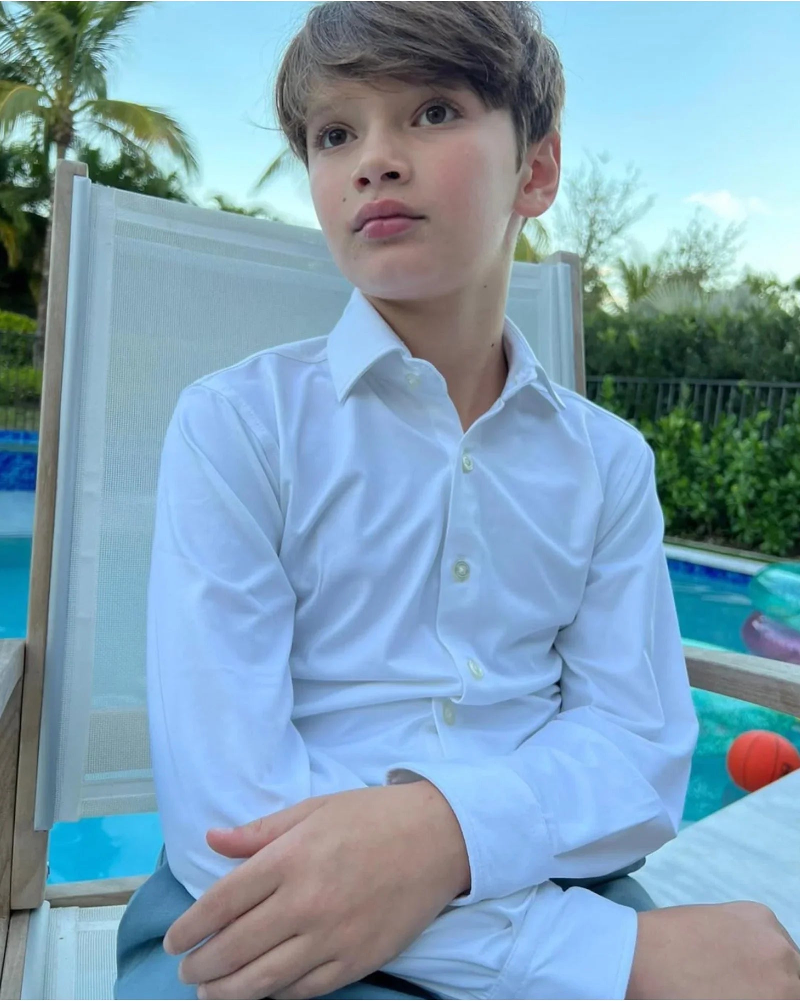 Child wearing a white shirt sitting by a pool with greenery in the background