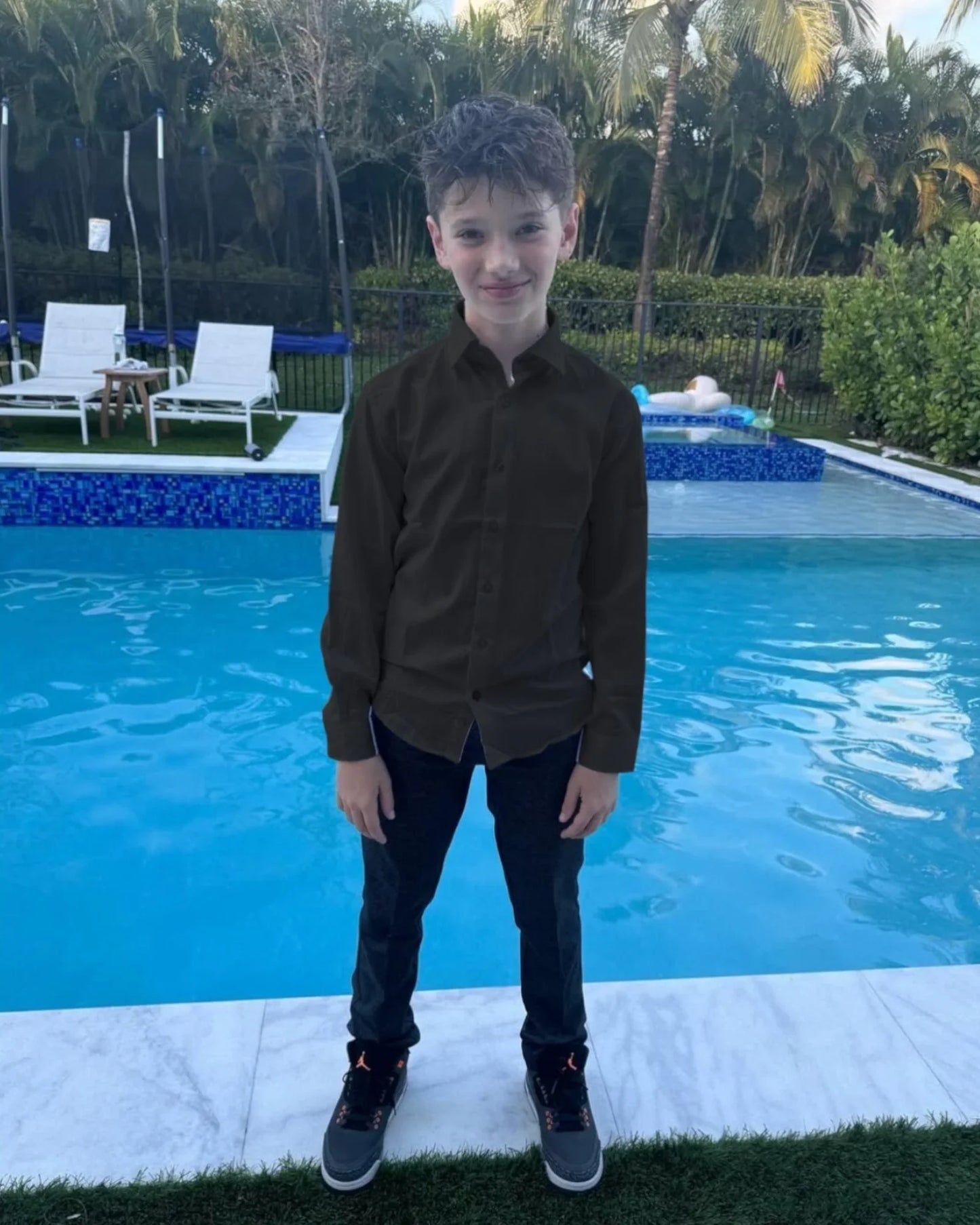 Young boy standing by a pool with greenery in the background, wearing black dress shirt
