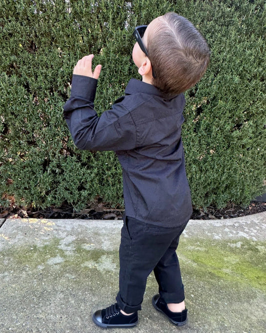 two year old boy wearing a black shirt and pants standing outdoors with greenery in the background