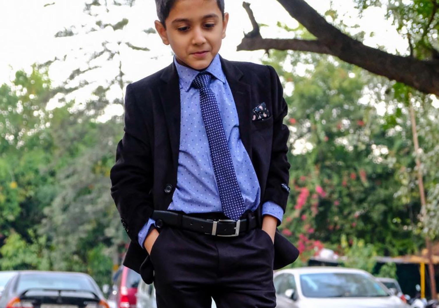 Young boy in formal attire standing on a street with cars and trees in the background