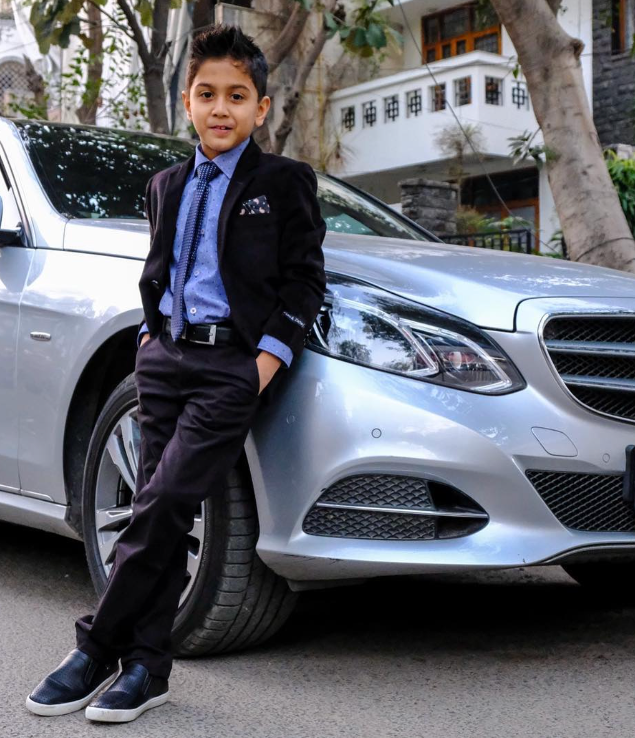 Child in formal attire leaning against a silver Mercedes-Benz car on a street.