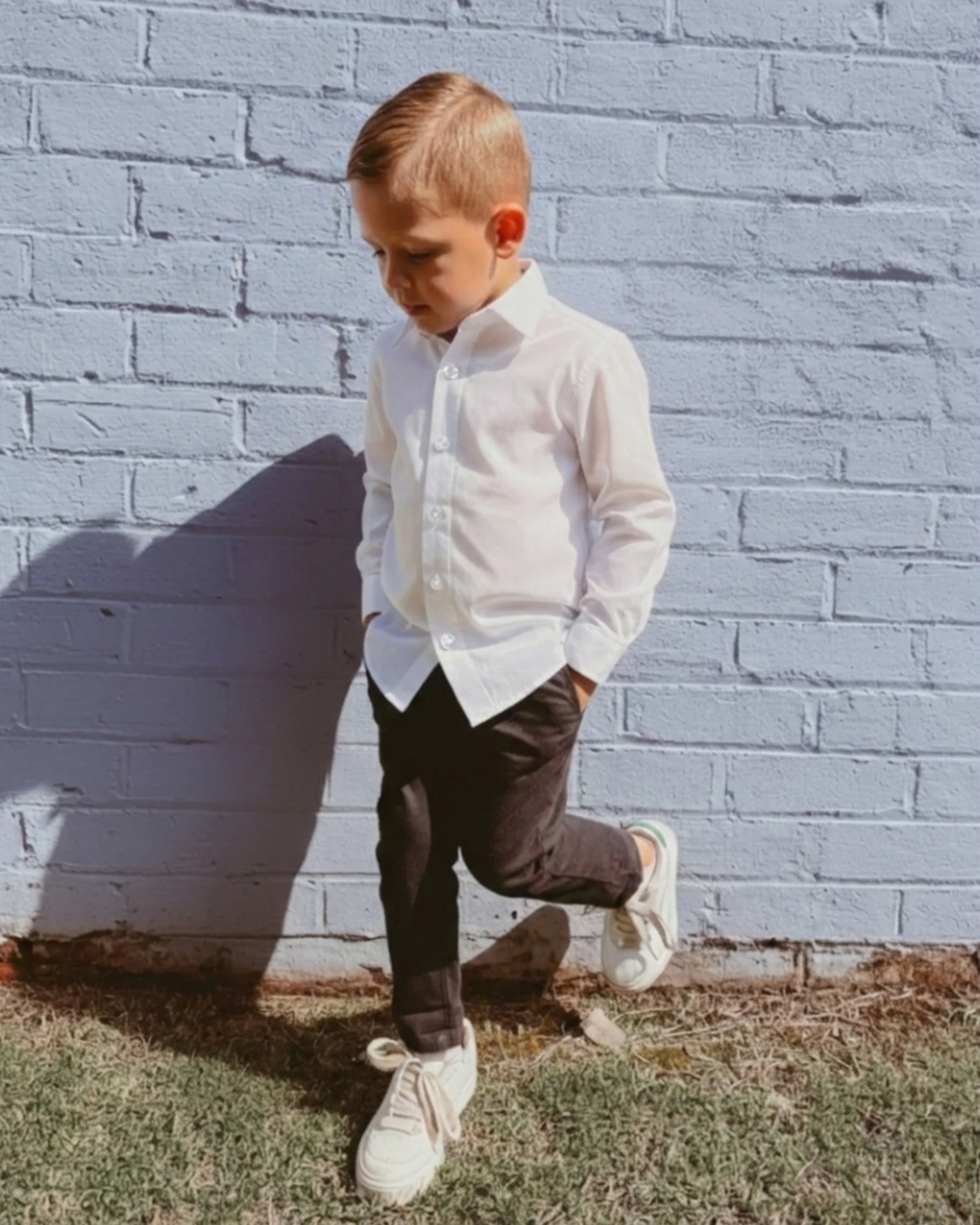Boy wearing a white shirt and black chinos standing against a brick wall