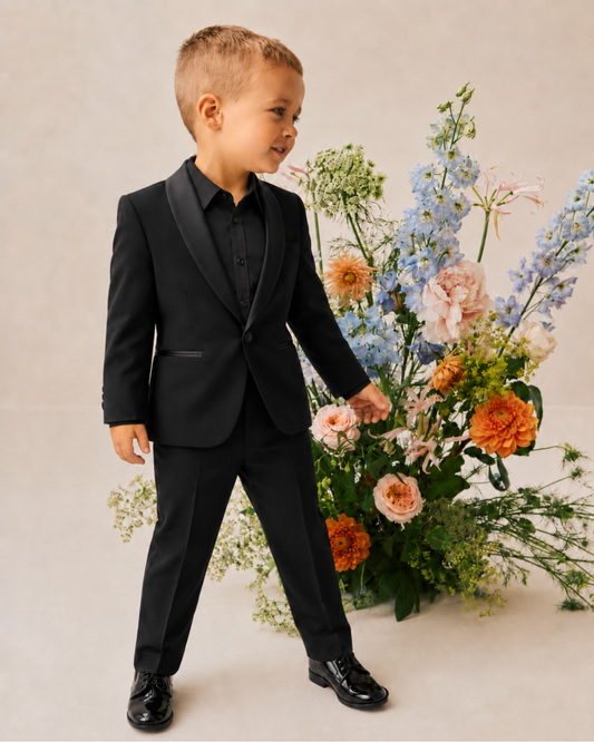 A young boy wearing a black boys' tuxedo suit with satin lapels, a black shirt, and black dress shoes, standing next to a large floral arrangement.