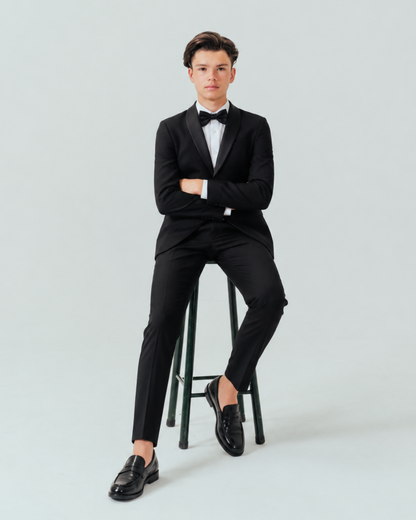 Teenager wearing a black tuxedo sitting on a stool against a plain background