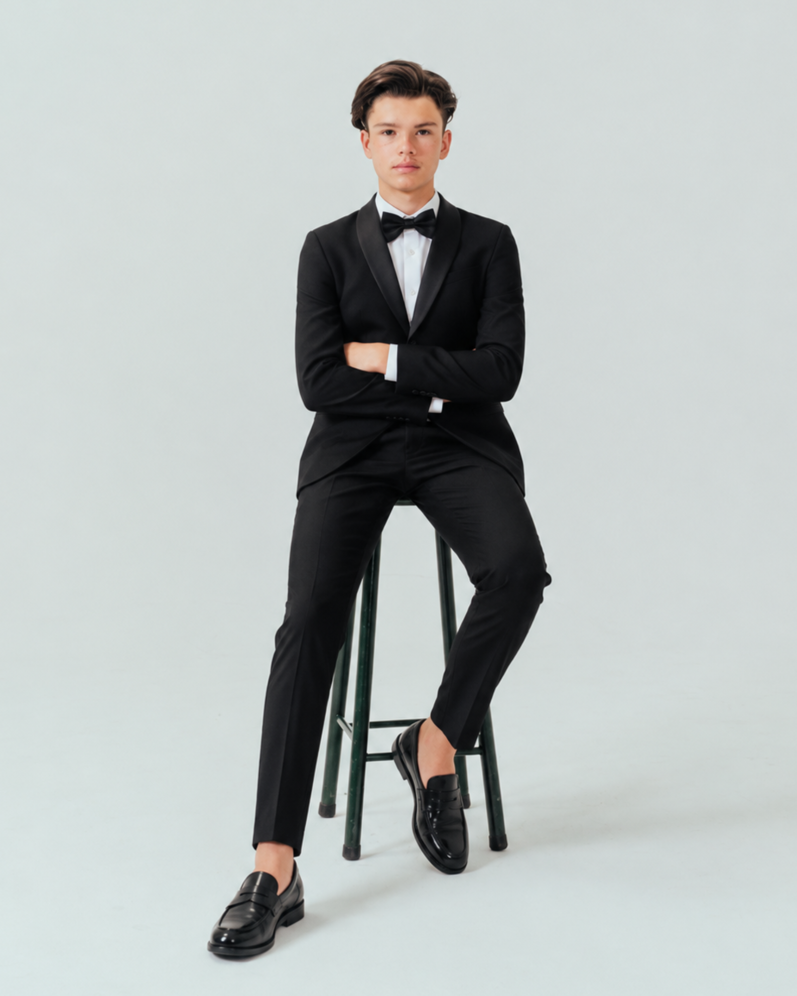 Teenager wearing a black tuxedo sitting on a stool against a plain background