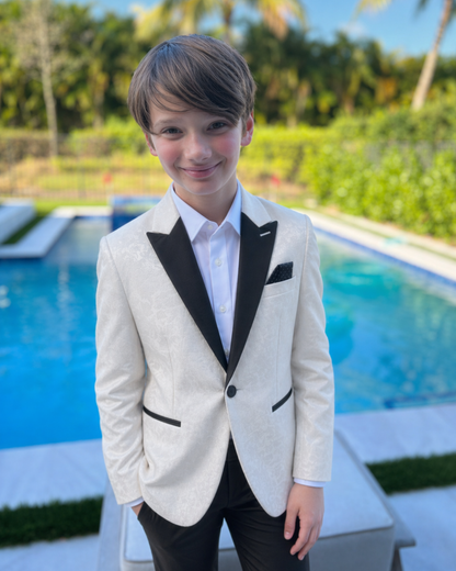 Young boy in a formal white blazer with black lapels standing by a pool.