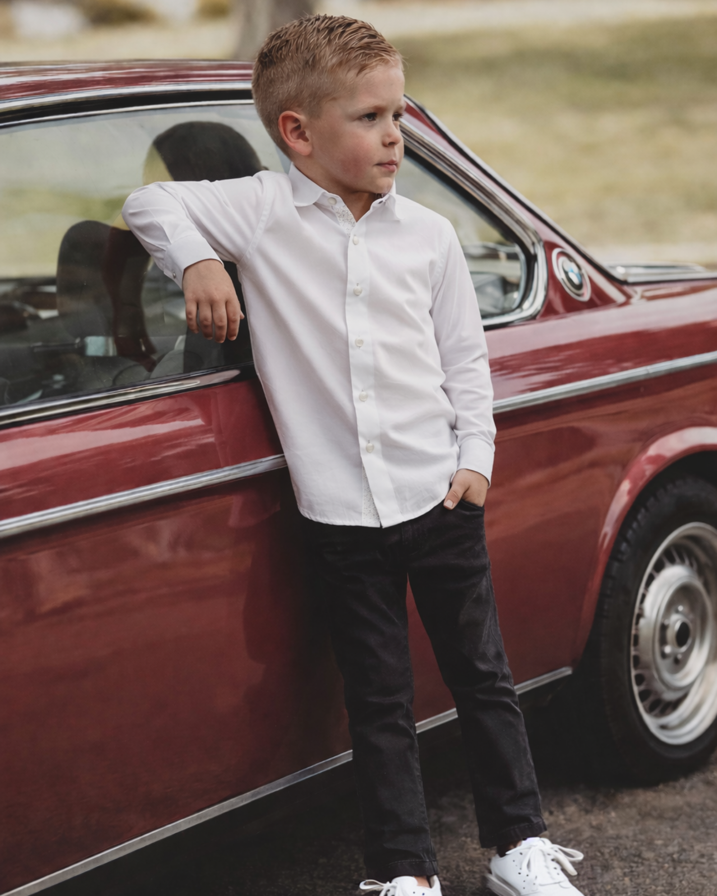 Young boy in a white shirt and black chinos leaning against a red vintage car.