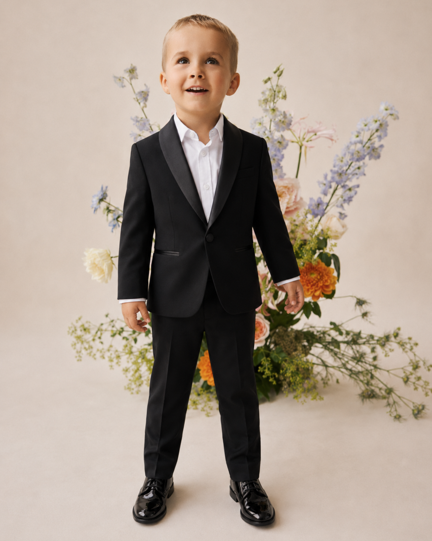 Young boy in a black suit standing in front of floral decorations.