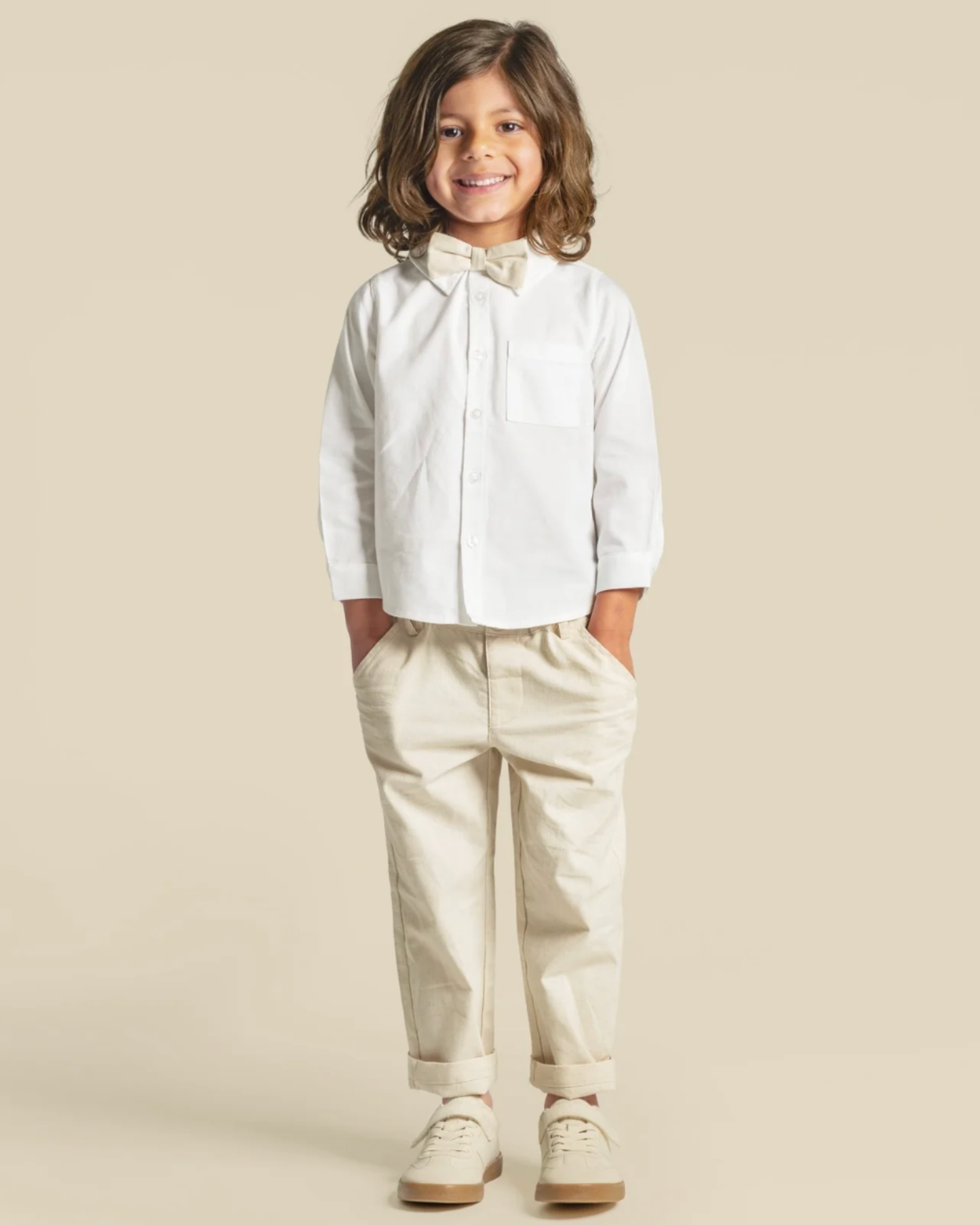 A young child modeling a white long-sleeve button-up shirt with a matching bow tie, paired with beige pants and white sneakers, set against a solid beige background.