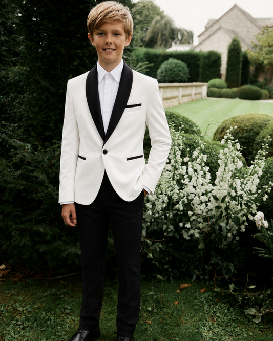 Young boy in a white suit with black lapels, with a white shirt and black dress pants standing outdoors in a garden.