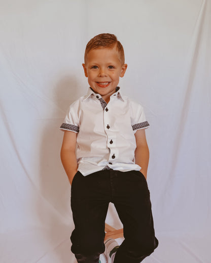 Young boy wearing a white shirt with black buttons and black pants against a plain background
