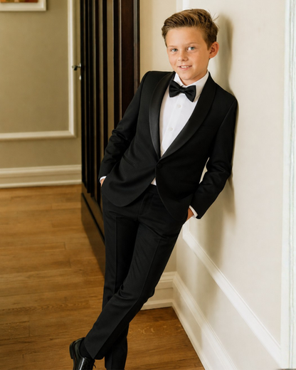 Boy in a black tuxedo with satin shawl lapel and bow tie, leaning against a wall.