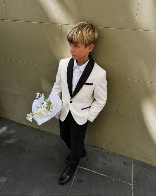 Young boy standing against a wall, wearing a white velvet blazer and white shirt and black dress pants, holding a bouquet of flowers 