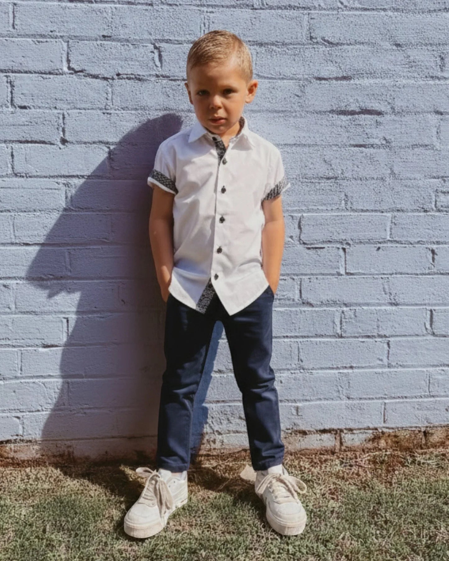 Young boy standing against a light blue brick wall wearing a white shirt and navy blue chinos.