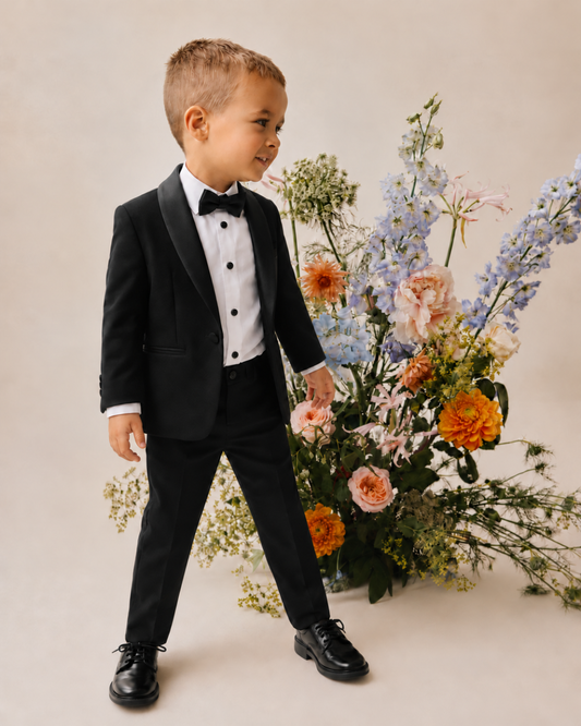 Young boy wearing a black tuxedo with satin lapels, a white dress shirt and black bow tie, standing beside pastel flowers.