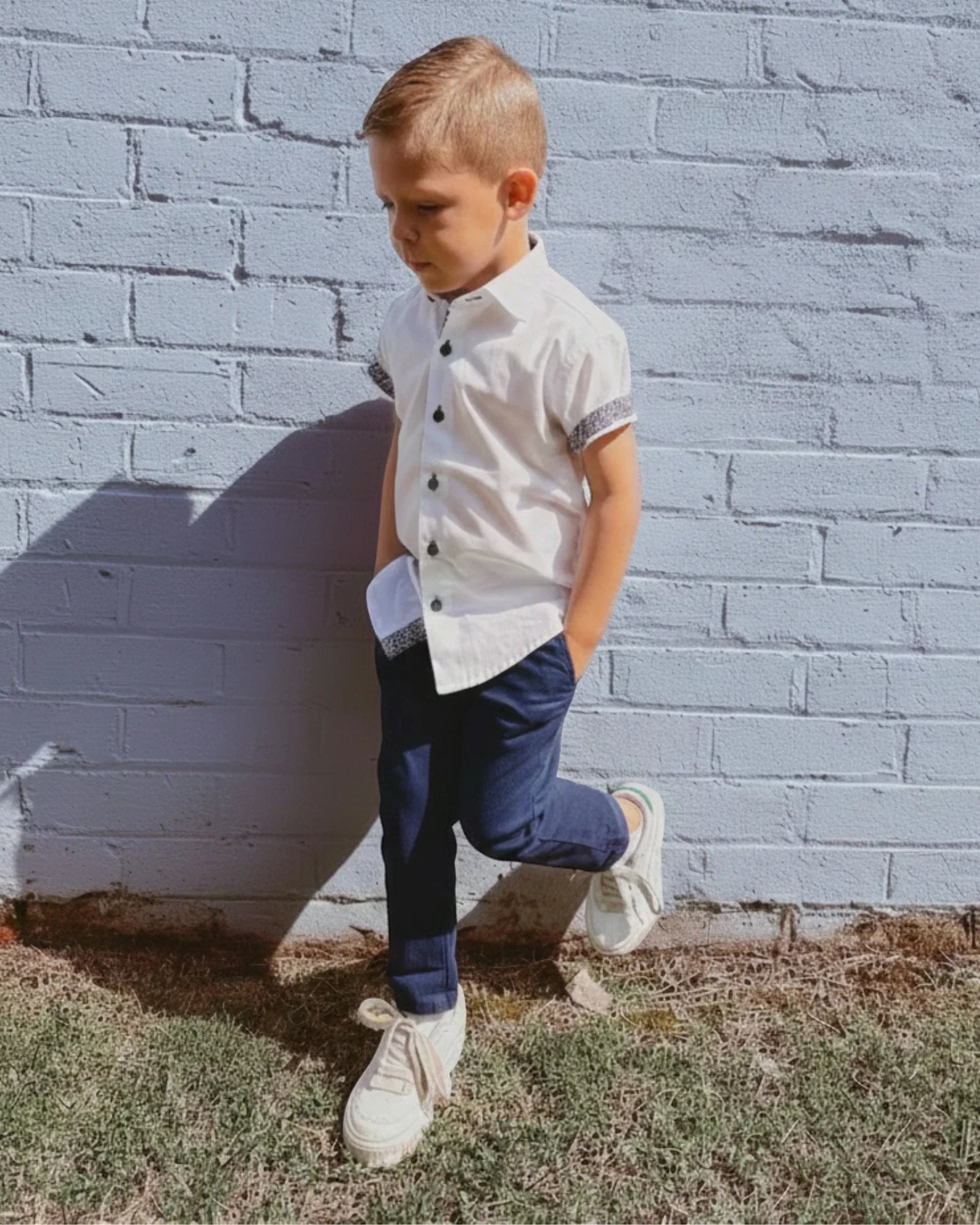 Young boy in a white shirt and navy blue chino pants standing against a gray brick wall.
