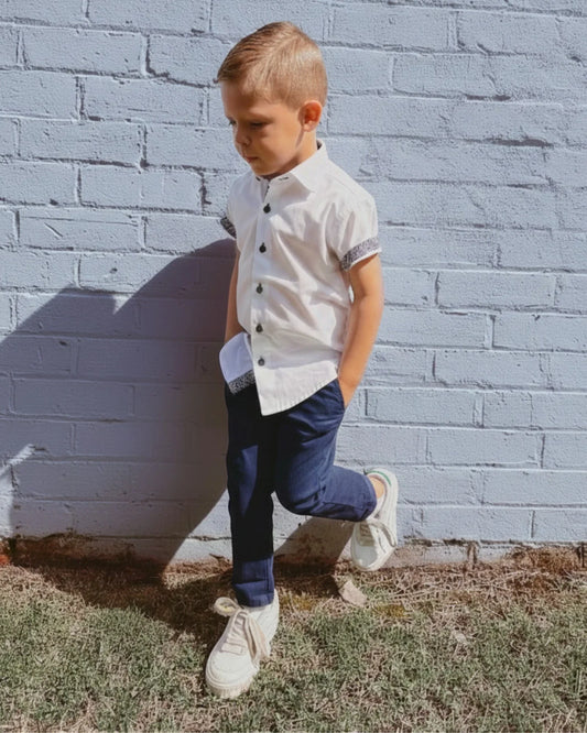Young boy in a white shirt and navy blue chino pants standing against a gray brick wall.
