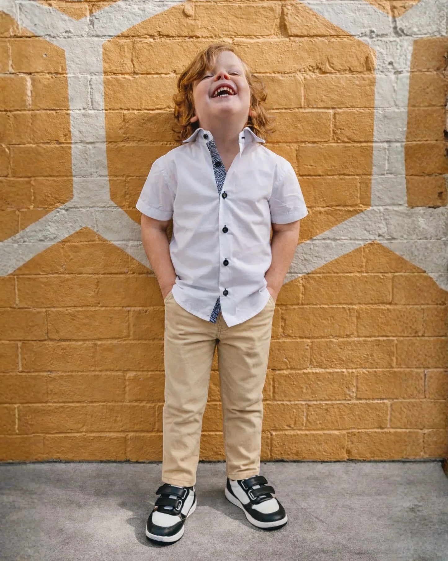 Child standing against a wall wearing a white shirt, beige chinos, and black shoes.