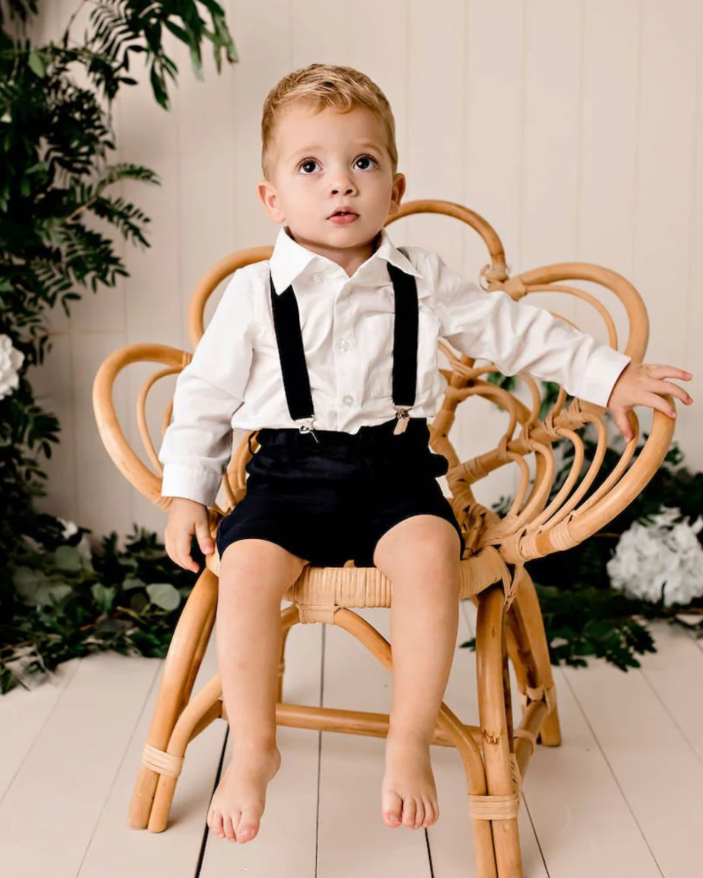A toddler boy sitting on a chair wearing a white long-sleeve button-up shirt, black suspenders, and black shorts.