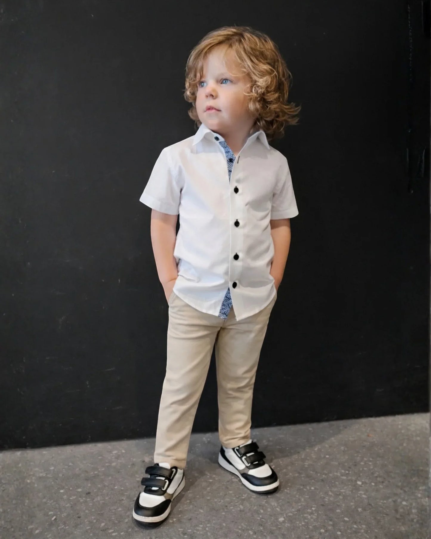 Child wearing a white shirt, beige chinos, and black and white shoes against a black background.