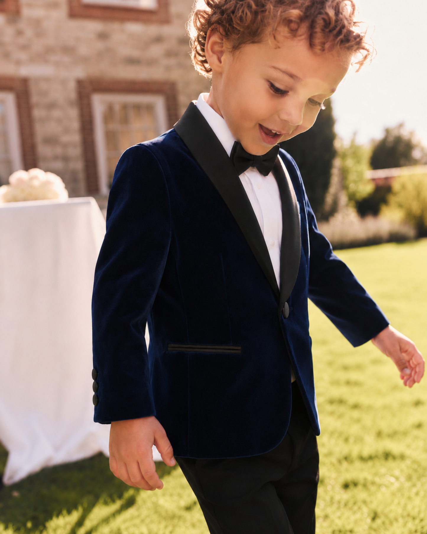 A young boy wearing a navy blue velvet tuxedo jacket with black satin lapels, a white dress shirt, and a black bow tie, walking outdoors on a green lawn.