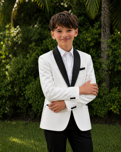 Young boy in a white tuxedo with black lapels standing outdoors with greenery in the background