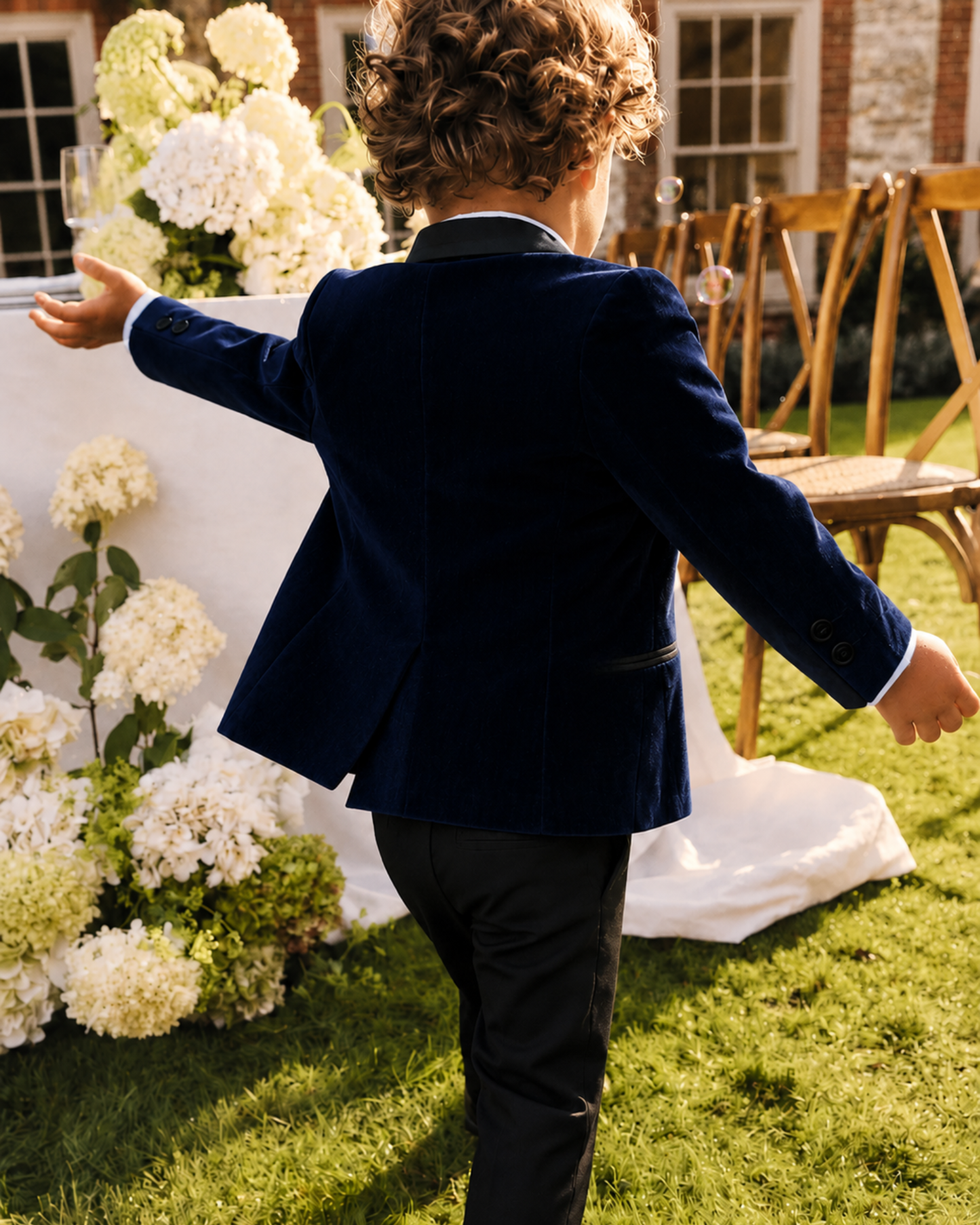 Back view of a boy wearing a boys navy velvet blazer with a black satin shawl lapel and black formal pants at an outdoor wedding.