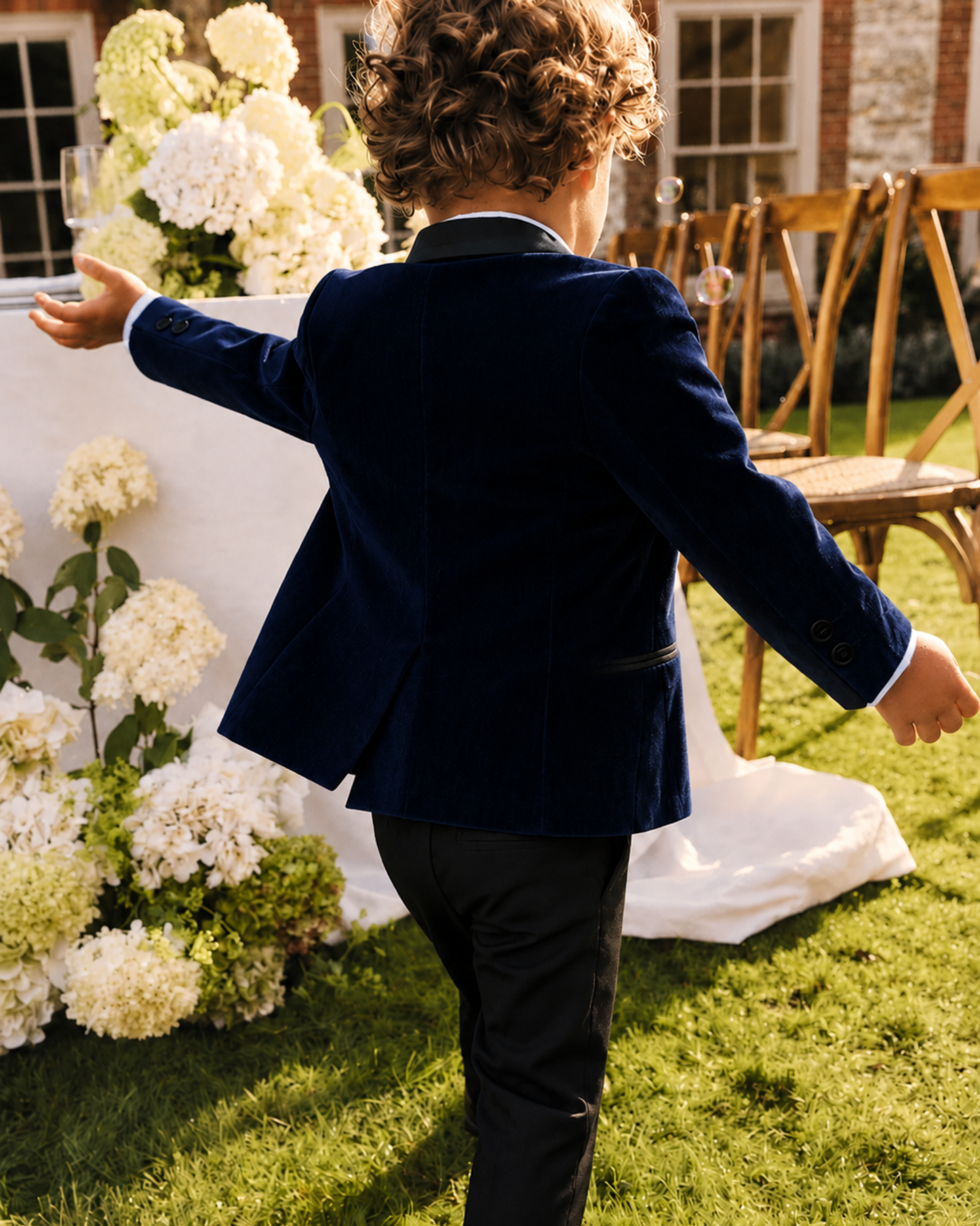 Back view of a boy wearing a boys navy velvet blazer with a black satin shawl lapel and black formal pants at an outdoor wedding.