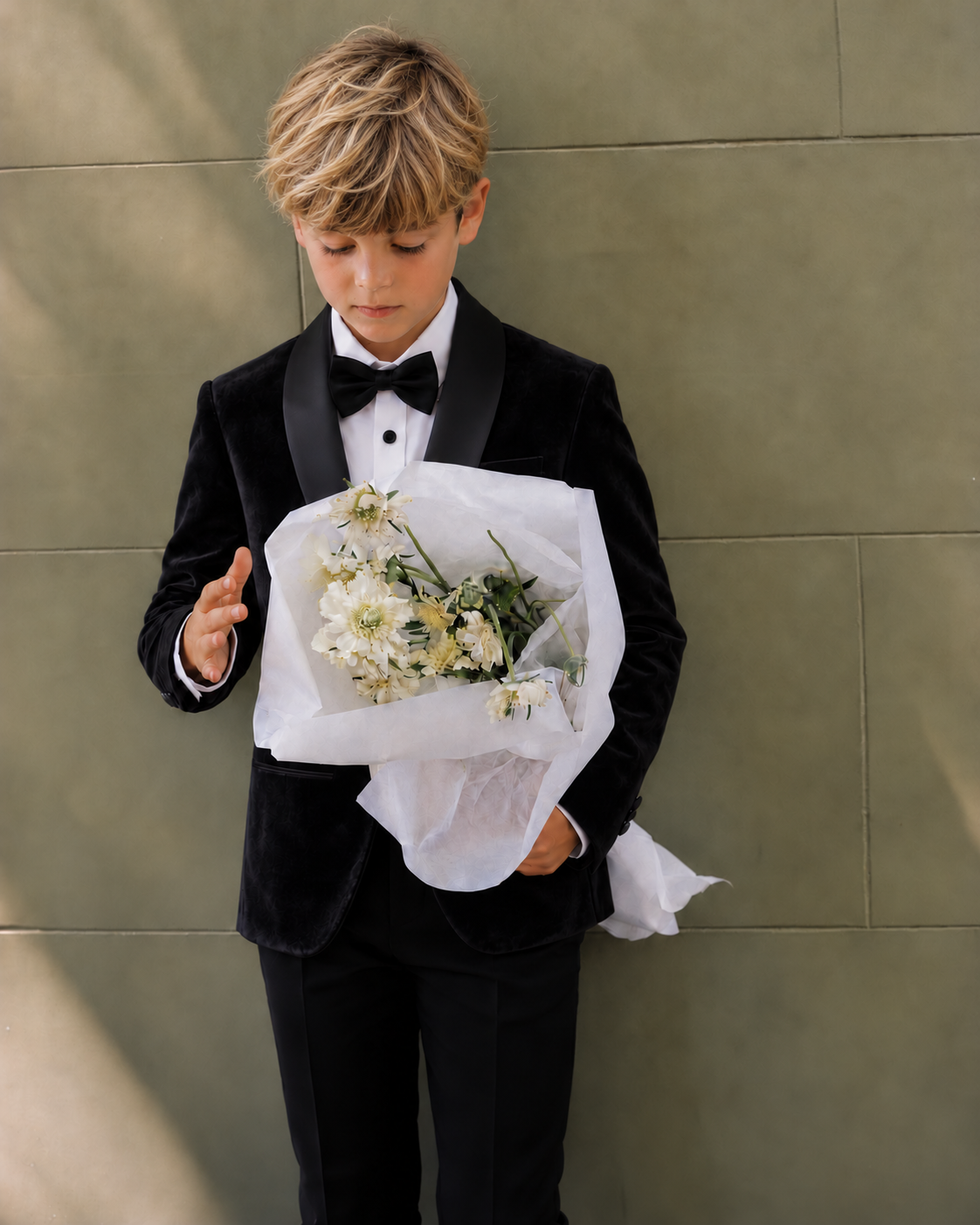 A young boy wearing a black velvet tuxedo jacket with black satin lapels, a white dress shirt, and a black bow tie, holding a bouquet of white flowers against a textured olive-green wall.