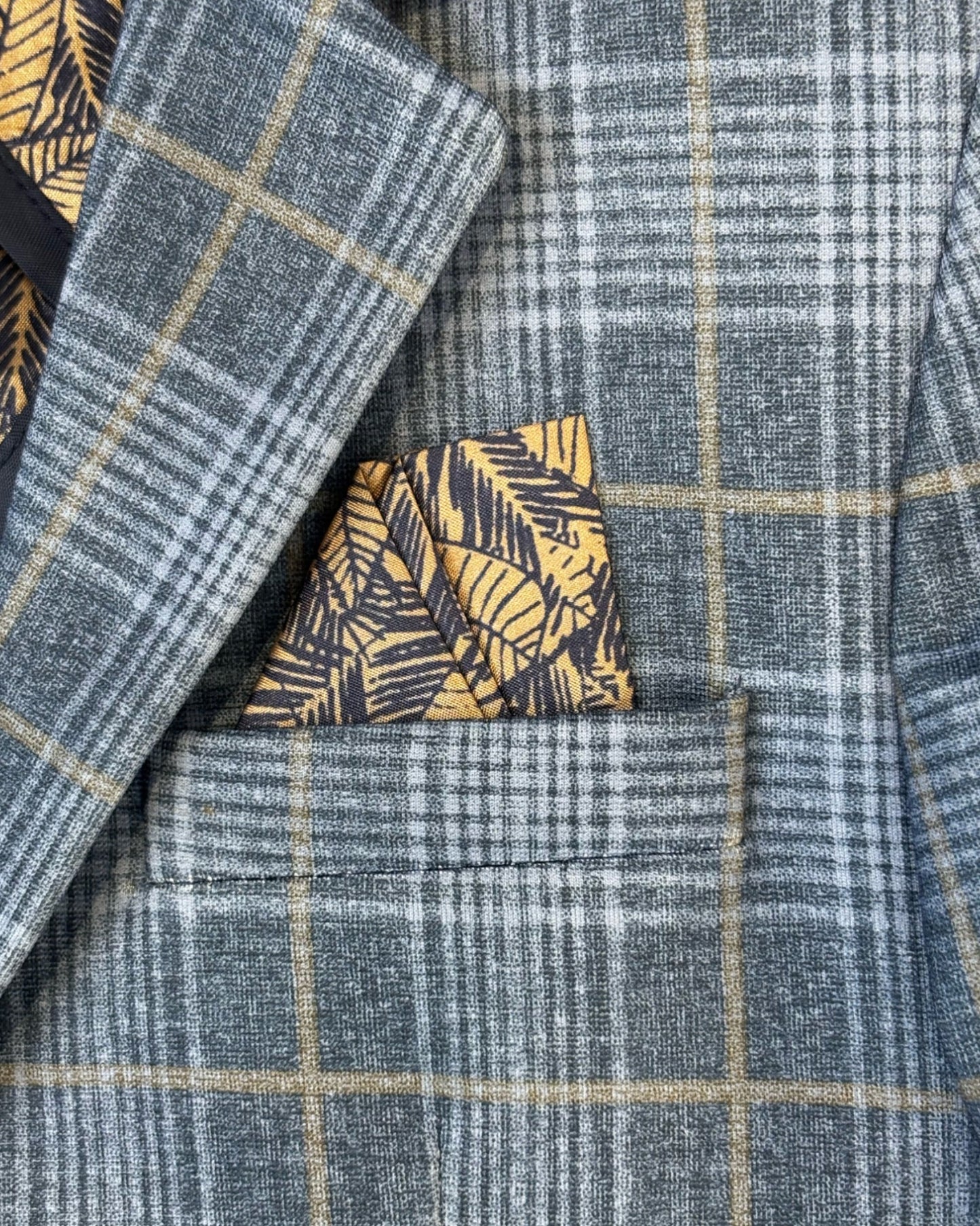 Close-up of a blue and grey plaid blazer with yellow windowpane lines, featuring a folded yellow and navy patterned pocket square tucked into the chest pocket.