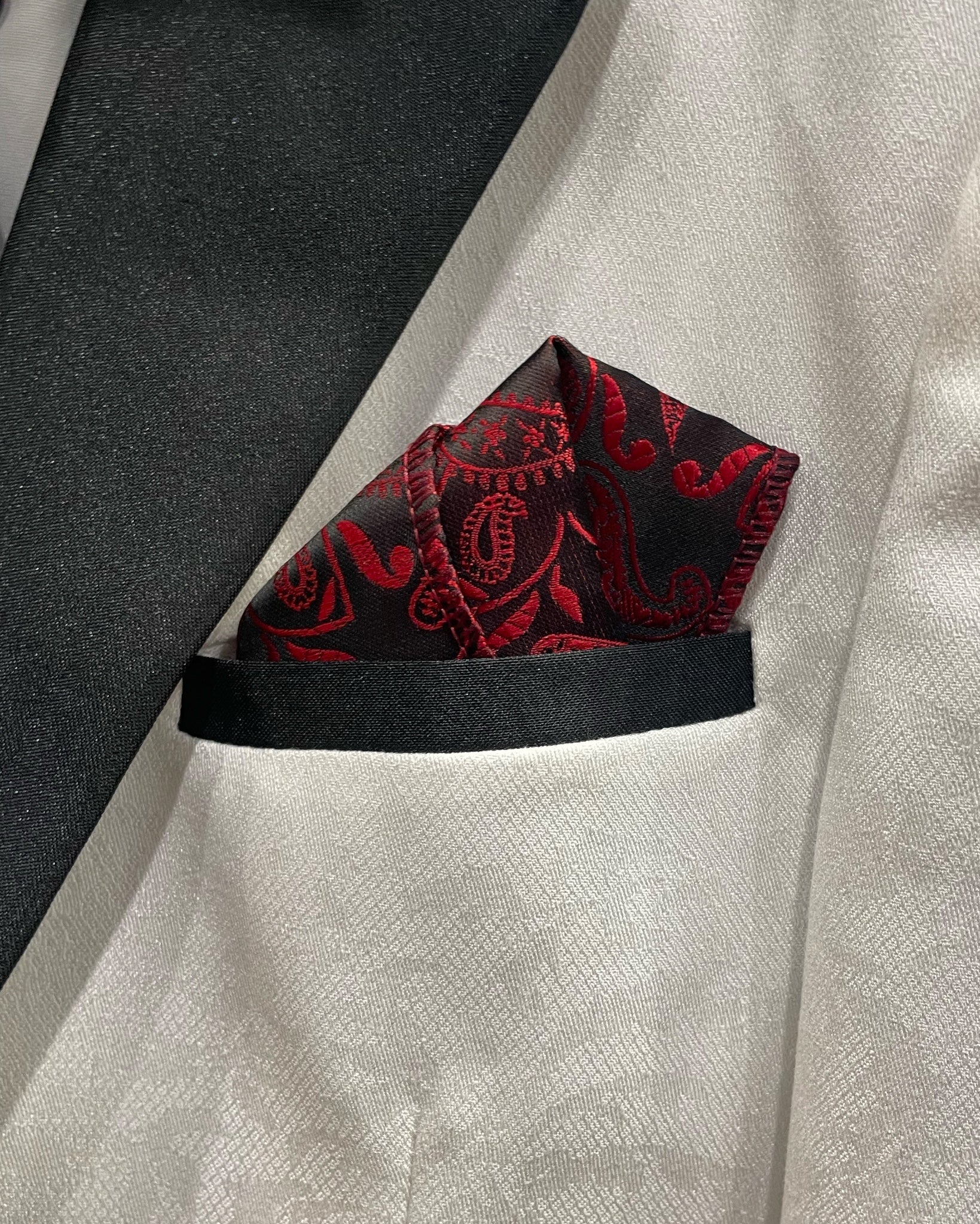 A close-up of a white jacquard suit jacket with a black lapel, featuring a red and black paisley patterned pocket square tucked into the breast pocket.