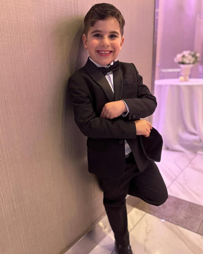 boy wearing black tux, including black shawl lapel blazer, white shirt, black dress pants, and black bow tie. he's standing against a wall and smiling.