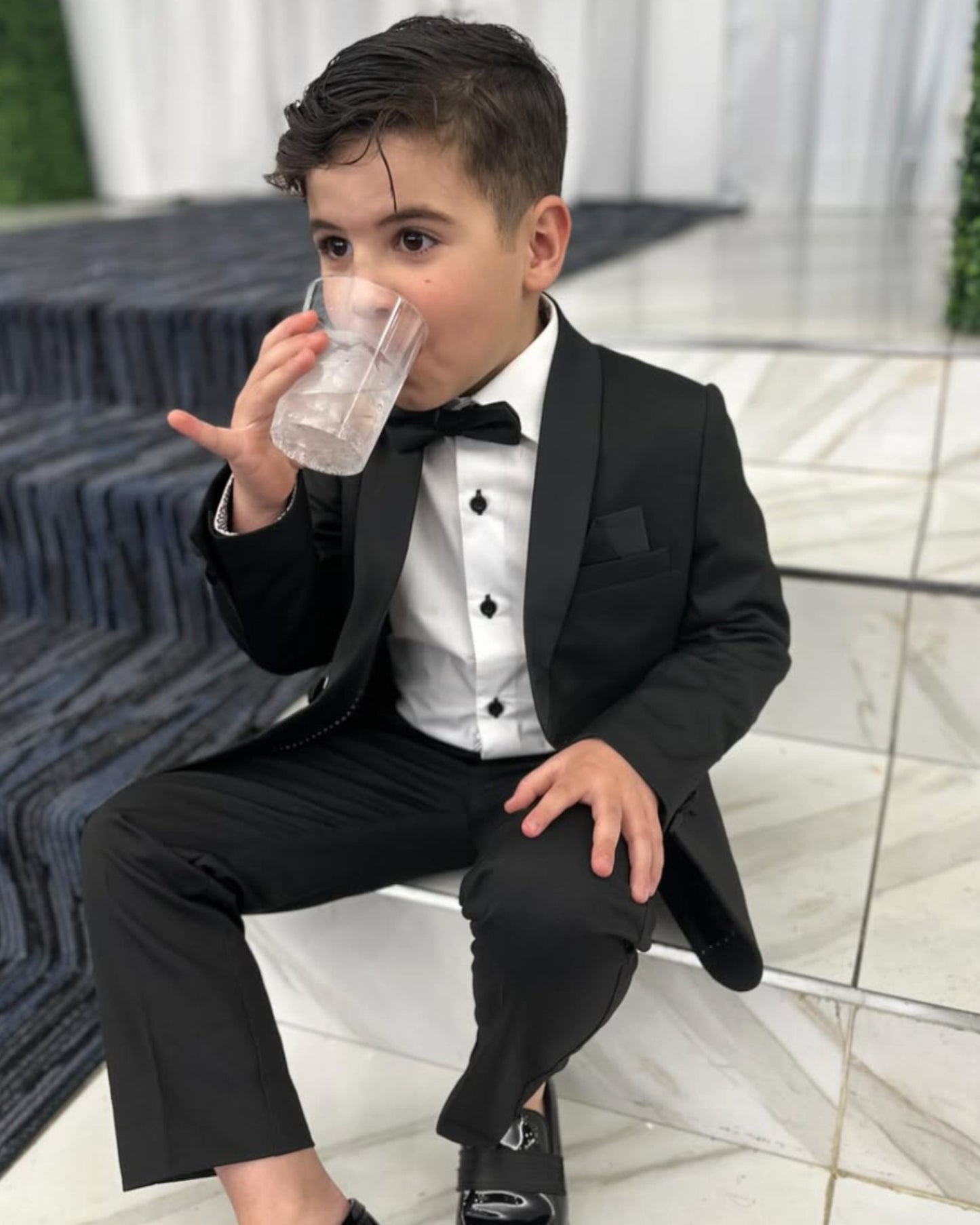boy sitting down and wearing black tux, including black shawl lapel blazer, white shirt, black dress pants, and black bow tie.
