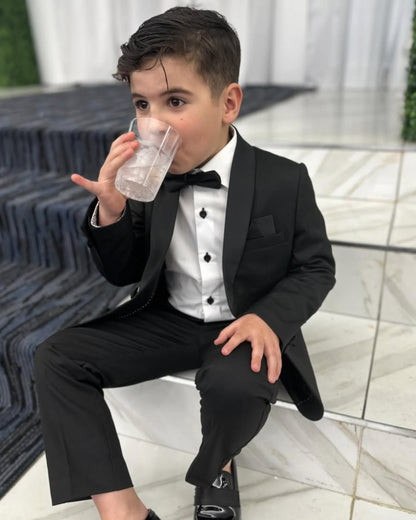 boy sitting down and wearing black tux, including black shawl lapel blazer, white shirt, black dress pants, and black bow tie.