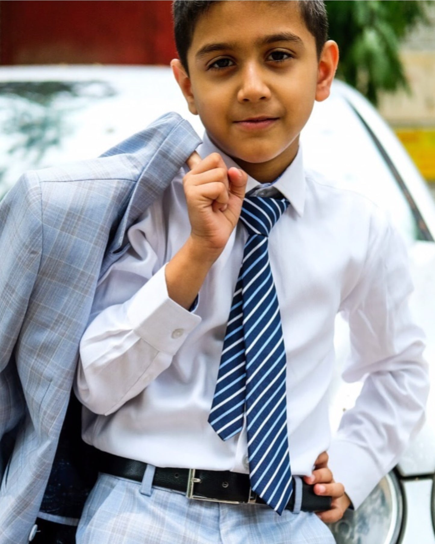 Young boy dressed in formal attire, including white dress shirt, striped blue tie, check pants, black belt, and sky blue checkered blazer. He is standing in front if a car.