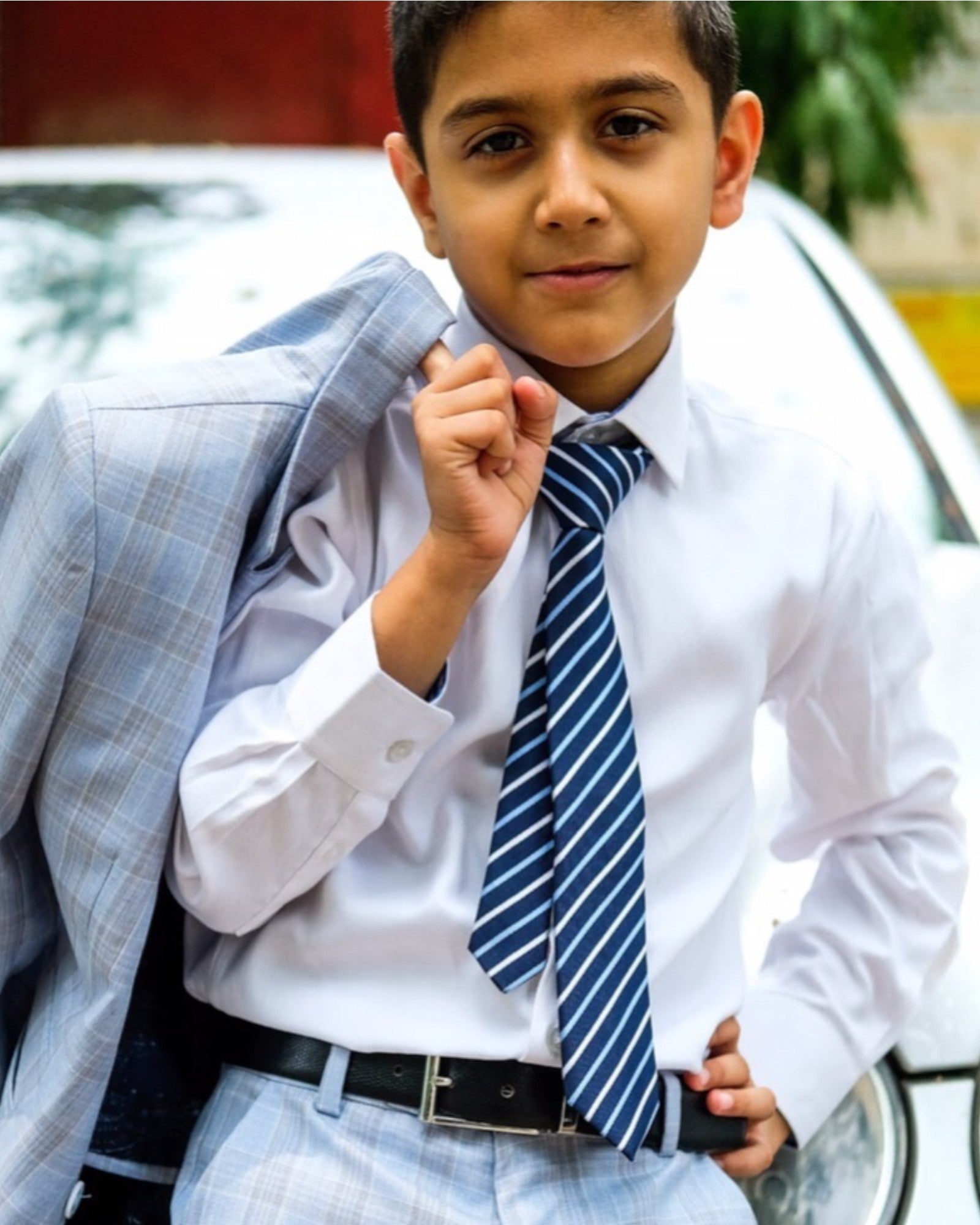 Young boy dressed in formal attire, including white dress shirt, striped blue tie, check pants, black belt, and sky blue checkered blazer. He is standing in front if a car.
