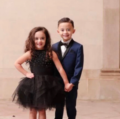 Two children, a girl in a black dress and a boy in a suit, standing against a beige wall.