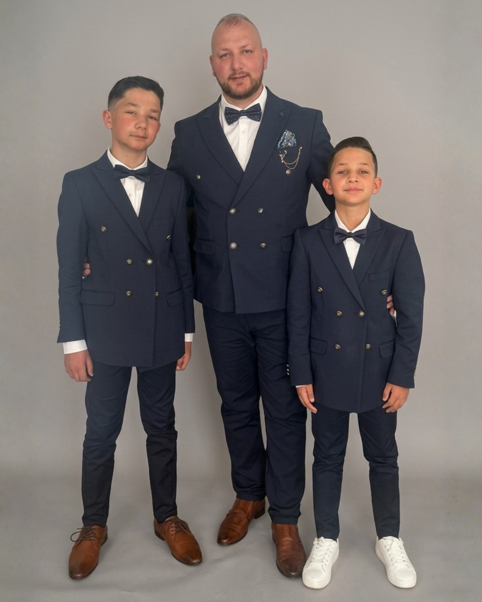 Man and two boys standing together in a studio, wearing matching navy blue double breasted suits with white shirts and bow ties, facing the camera against a plain light grey background