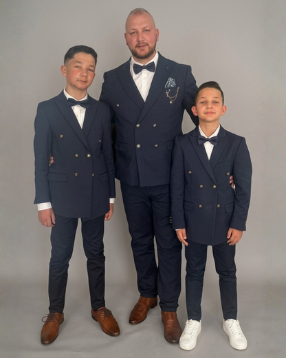 Man and two boys standing together in a studio, wearing matching navy blue double breasted suits with white shirts and bow ties, facing the camera against a plain light grey background