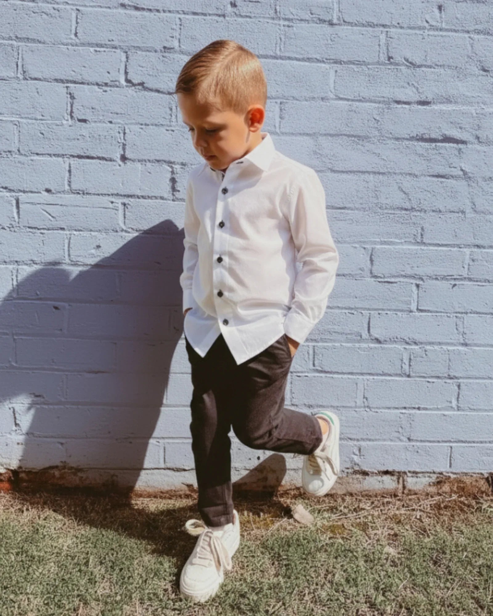 Young boy in a white shirt and black chinos standing against a light blue brick wall.