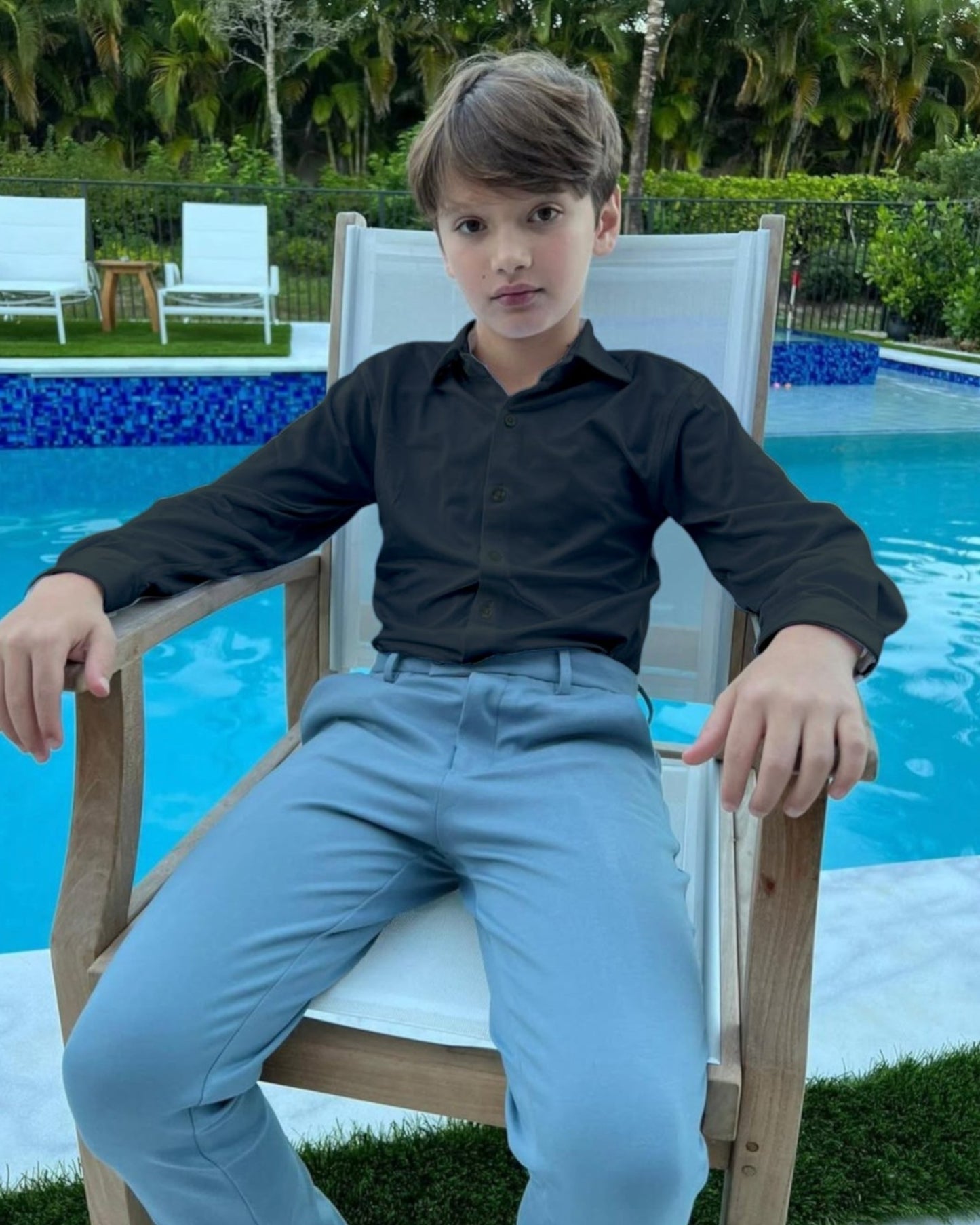 Young boy sitting on a chair by a pool with greenery in the background, wearing black dress shirt