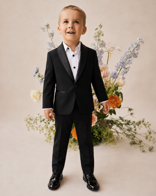 Young boy wearing a black tuxedo with satin lapels, a white shirt with black buttons and black dress shoes, standing in front of flowers.