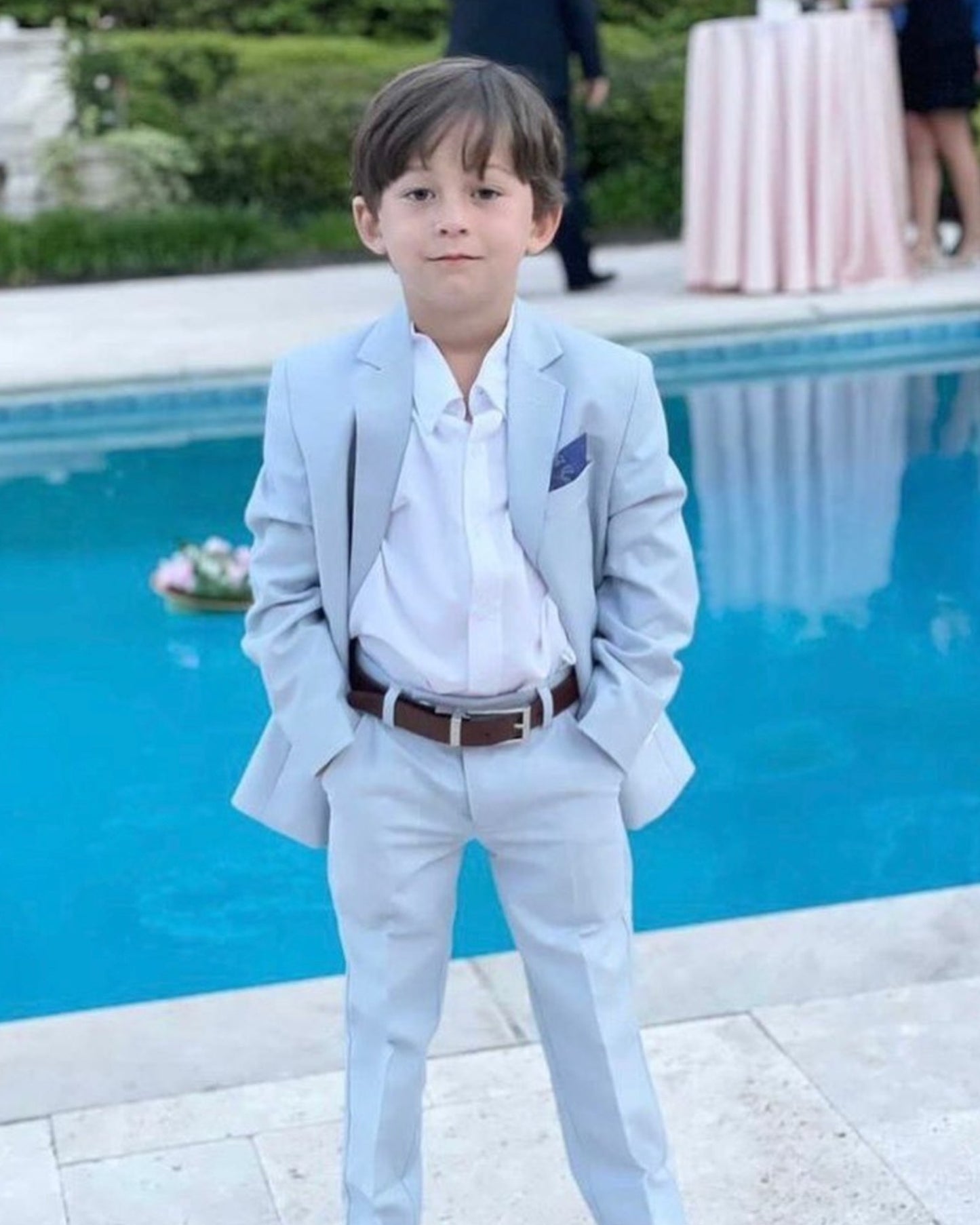 Young boy in a light grey suit standing by a pool. with white dress shirt being featured. 
