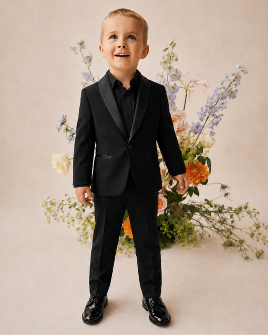 A young boy wearing a black formal tuxedo with a black shirt and shiny black dress shoes, standing in front of a beige background with a floral arrangement.